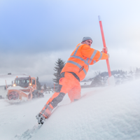 Ein Winterdienst-Mitarbeiter setzt ein Schneezeichen an den Straßenrand in verschneiter Landschaft. Im Hintergrund ein Schneepflug. 