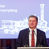 Verkehrsminister Christian Bernreiter steht an einem Rednerpult und hält eine Rede. Im Hintergrund eine Leinwand mit Aufdruck der Adler-Lok und Text „Bayerischer Eisenbahnempfang 2022“.